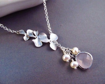 May include: A silver necklace featuring a floral design with three detailed flower pendants. The necklace includes a cluster of small pearls and a faceted, light pink gemstone pendant. The chain is delicate and the overall design is elegant.