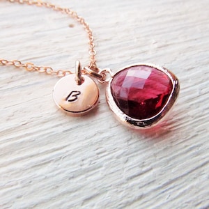 May include: A rose gold necklace with a small round pendant engraved with the letter "B" and a larger teardrop-shaped pendant with a red gemstone.