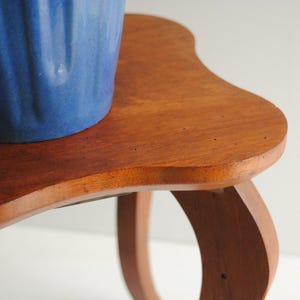 K&ouml;nnte beinhalten: Kleiner Beistelltisch aus hellem Holz mit geschwungener Tischplatte und Beinen. Auf dem Tisch steht ein blauer Keramiktopf mit strukturierter Oberfl&auml;che. Das Holz ist hellbraun.