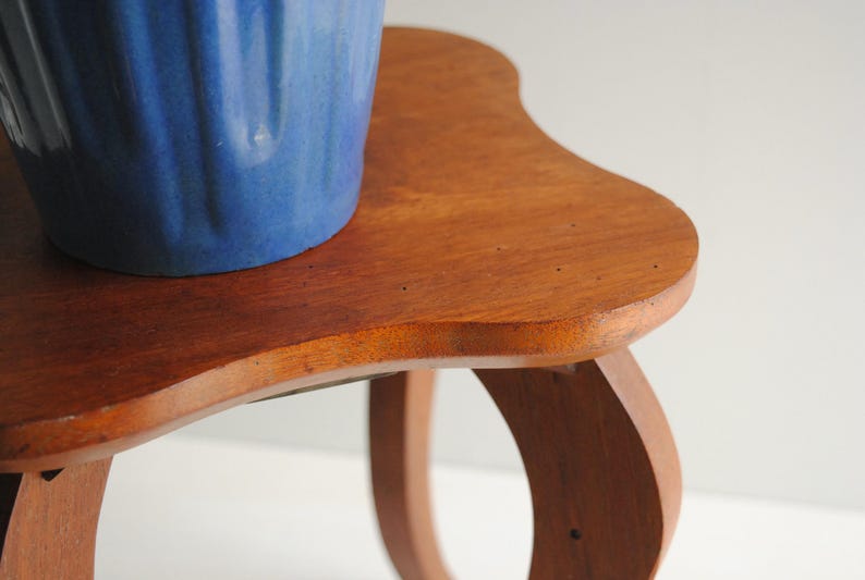 K&ouml;nnte beinhalten: Kleiner Beistelltisch aus hellem Holz mit geschwungener Tischplatte und Beinen. Auf dem Tisch steht ein blauer Keramiktopf mit strukturierter Oberfl&auml;che. Das Holz ist hellbraun.