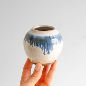 Pode incluir: Pequeno vaso redondo de cerâmica com base creme e esmalte azul e azul escuro que escorre pelas laterais. O vaso tem uma superfície texturizada e uma pequena abertura no topo.