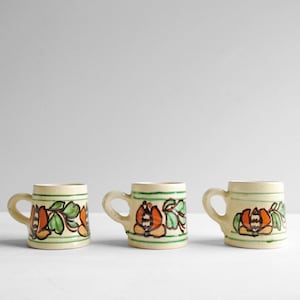 May include: Three small ceramic mugs with a cream-colored base, each featuring a hand-painted design of orange and brown flowers with green leaves, accented by two horizontal green stripes encircling the top and bottom of the mug body.