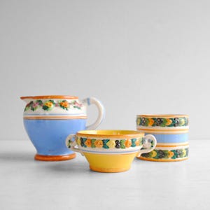 May include: Three pieces of vintage ceramic tableware. A blue pitcher with a white handle and floral trim, a yellow bowl with handles and floral trim, and a blue and white cylindrical container with floral trim.