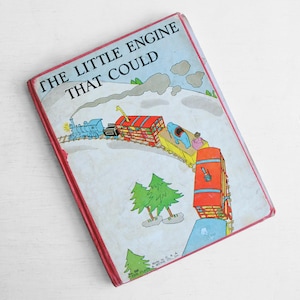 Könnte beinhalten: Vintage Kinderbuch mit dem Titel "The Little Engine That Could". Das Cover zeigt eine farbenfrohe Illustration einer blauen Lok, die verschiedene Waggons zieht, darunter einen roten und einen gelben. Das Buch hat einen roten Rand.