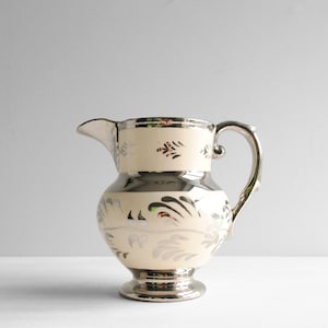 May include: A ceramic pitcher featuring a cream-colored base with metallic silver accents. The design includes a silver-finished handle, rim, and base, complemented by repeating silver leaf-like patterns encircling the body and neck of the vessel.