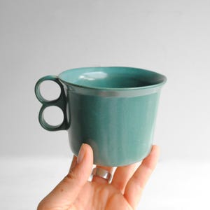 May include: A teal ceramic mug with a smooth, matte finish and a unique handle design featuring two stacked circular loops. The mug has a wide, open top and a slightly tapered body, showcasing a solid, consistent color throughout the entire piece.
