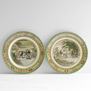 May include: Two round ceramic plates featuring detailed, colorful illustrations. The left plate depicts an indoor tavern scene with figures, while the right plate shows an outdoor landscape with a person riding a horse. Both have ornate borders.