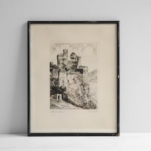 May include: A framed black and white etching depicting a stone castle perched on a cliffside overlooking a body of water. The artwork features detailed architectural lines, trees, and a rocky landscape, enclosed in a simple black frame.