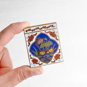Pode incluir: Um estojo compacto retangular de metal com esmalte branco e detalhes florais vermelhos. O centro exibe um painel azul em forma de amêndoa com uma cena colorida de dois patos nadando entre flores de lótus e vitórias-régias verdes, contornado em metal dourado.