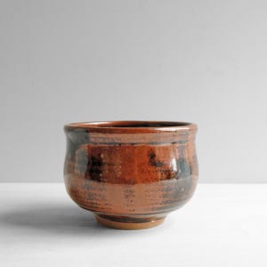 May include: A small, rounded ceramic bowl featuring a glossy, variegated glaze in shades of deep burnt orange, copper, and dark brown. The bowl has a wide, open mouth, a slightly curved body, and a narrow, circular base, showcasing a smooth finish.
