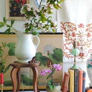 K&ouml;nnte beinhalten: Ein Stillleben mit einer wei&szlig;en Kanne, einem kleinen Holzschemel und einem gerahmten Blumenbild. Eine kleine braune Pferdfigur sitzt auf dem Schemel. Eine gr&uuml;ne Lampe und eine Topf-Orchidee sind ebenfalls vorhanden.