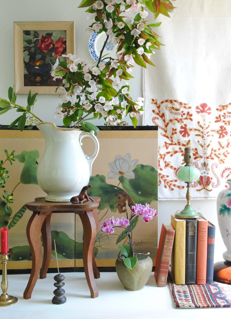 K&ouml;nnte beinhalten: Ein Stillleben mit einer wei&szlig;en Kanne, einem kleinen Holzschemel und einem gerahmten Blumenbild. Eine kleine braune Pferdfigur sitzt auf dem Schemel. Eine gr&uuml;ne Lampe und eine Topf-Orchidee sind ebenfalls vorhanden.