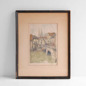 Op de afbeelding: Een ingelijste prent van een Europees stadsgezicht. Het kunstwerk toont gebouwen langs een rivier, een stenen brug en een kathedraal op de achtergrond. De zwarte lijst heeft een verweerde uitstraling.