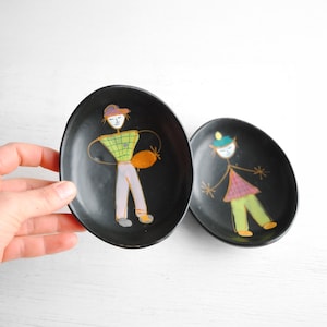 Op de afbeelding: Twee zwarte keramische sierborden met handgeschilderde figuren. Een bord heeft een figuur met een groen-geel geruit overhemd, een paarse broek en een hoed. Het andere bord heeft een figuur met een roze-paars geruit overhemd, een groene broek en een hoed.