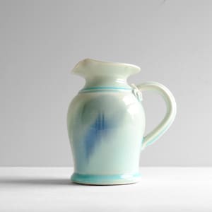 May include: A ceramic pitcher featuring a glossy light blue and white glaze with a darker blue abstract wash pattern. The vessel has a curved handle, a flared rim with a pouring spout, and a defined base, showcasing a smooth, reflective finish.