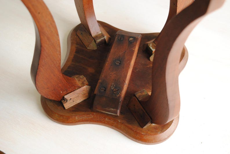 K&ouml;nnte beinhalten: Nahaufnahme der Unterseite eines kleinen, antiken Holztisches. Der Tisch hat geschwungene Beine und einen dekorativen, wellenf&ouml;rmigen Rand. Das Holz hat einen warmen, r&ouml;tlich-braunen Farbton mit sichtbarer Maserung und Altersspuren.