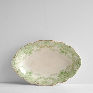 Puede incluir: Plato ovalado para servir con borde festoneado. El plato blanquecino presenta un diseño floral verde alrededor del borde y un ribete dorado. El centro del plato es de color blanquecino liso.