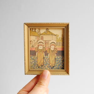 Puede incluir: Una ilustración vintage enmarcada de dos niñas con vestidos amarillos y gorros blancos de pie en un campo de flores. Las niñas están frente a un edificio con un techo a dos aguas.