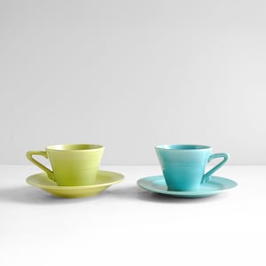 Puede incluir: Dos tazas de té y platillos de cerámica. Una es de color verde lima brillante, la otra turquesa vibrante. Ambas tienen un diseño clásico con asa y platillo a juego. Las tazas y los platillos están colocados sobre una superficie blanca.