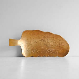 May include: A gold-colored, leaf-shaped tray with an etched floral design. The tray has a handle on one end and a scalloped edge. The floral pattern is centered on the tray and is surrounded by etched details.