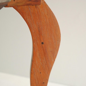 K&ouml;nnte beinhalten: Nahaufnahme eines h&ouml;lzernen Tischbeins mit geschwungenem Design. Das Bein besteht aus hellbraunem Holz und hat eine glatte, polierte Oberfl&auml;che. Das Bein ist an einer Tischplatte aus Holz befestigt. Der Hintergrund ist eine schlichte wei&szlig;e Wand.