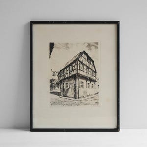 May include: A black and white etching print in a thin black frame depicting a corner view of a timber-framed building with a gabled roof and shuttered windows. The artwork features a cobblestone street and a hanging sign on the building exterior.