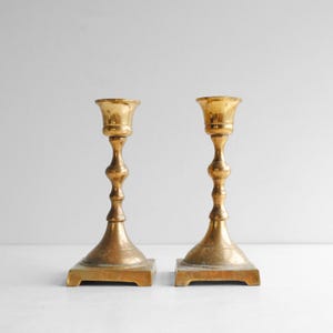 May include: Two antique brass candlesticks. Each candlestick has a square base, a decorative stem, and a flared top. The brass has a warm, aged patina. The candlesticks are displayed against a white background.
