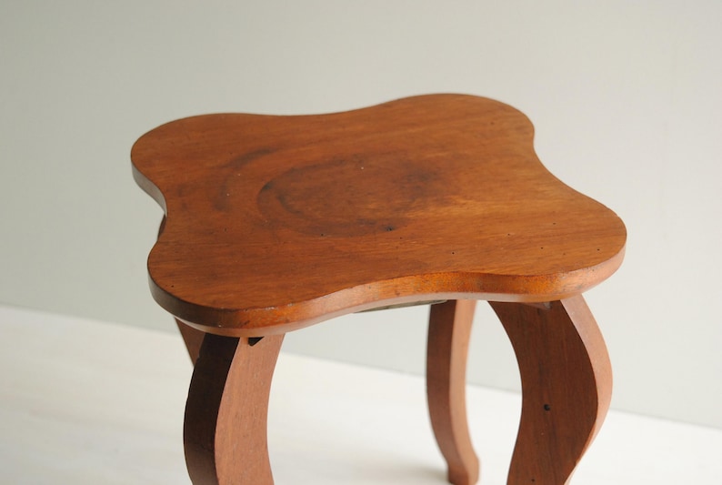 K&ouml;nnte beinhalten: Kleiner Holzschemel mit einer einzigartigen, geschwungenen Oberseite und Beinen. Der Schemel ist aus warmem, rotbraunem Holz gefertigt. Die Beine sind geschwungen und tragen zur Vintage-&Auml;sthetik des Schemels bei. Der Schemel ist etwa 30 cm hoch.