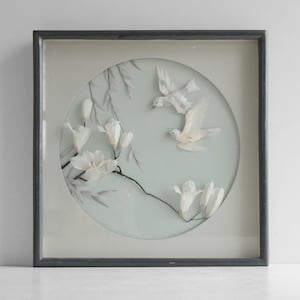 May include: Framed artwork featuring a circular design with white magnolia flowers and two white birds in flight. The artwork is set against a pale blue background and framed in a dark gray square frame.