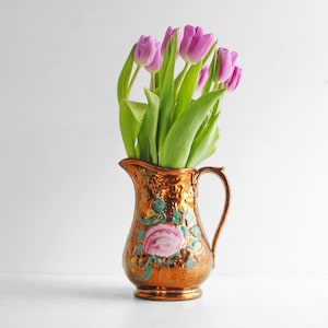 Può includere: Una brocca color rame con manico, piena di vivaci tulipani viola e foglie verdi. La brocca presenta un motivo floreale con una rosa rosa e accenti verdi e gialli. La brocca è un vaso decorativo.