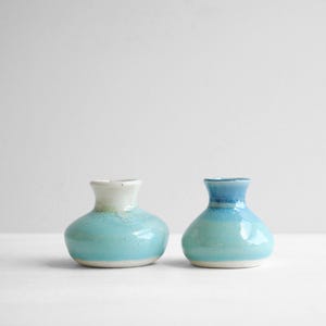 May include: Two small ceramic bud vases with a rounded base and a narrow neck. The left vase features a white upper section transitioning into a bright turquoise glaze. The right vase displays a blue upper section fading into a turquoise base.