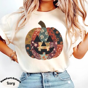 以下が含まれることがあります： アイボリー色のTシャツ。パッチワークのかぼちゃのデザイン。かぼちゃは黒い顔をしており、赤、緑、青の色合いの様々な花柄で構成されています。「Comfort Colors Ivory」の文字が下部にプリントされています。