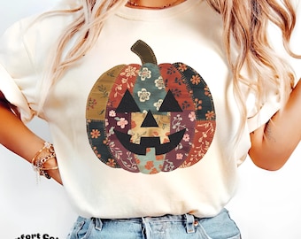 Puede incluir: Camiseta de color marfil con un diseño de calabaza patchwork. La calabaza tiene una cara negra y está compuesta por varios estampados florales en tonos rojos, verdes y azules. El texto "Comfort Colors Ivory" está impreso en la parte inferior.