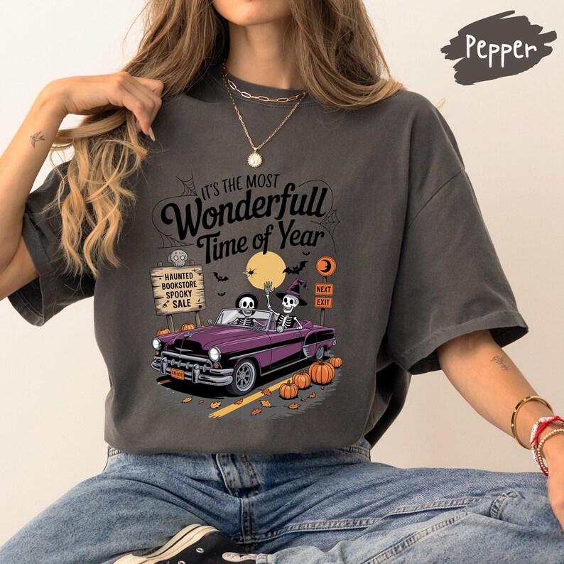 Puede incluir: Camiseta de color pimienta con un gr&aacute;fico de Halloween. El dise&ntilde;o presenta un coche morado con esqueletos al volante, calabazas y el texto "It's the Most Wonderfull Time of Year". Un letrero dice "Haunted Bookstore Spooky Sale".