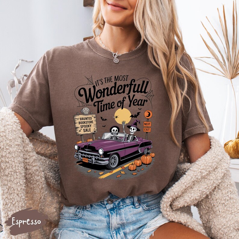 Puede incluir: Camiseta marr&oacute;n con un dise&ntilde;o de Halloween. El dise&ntilde;o presenta un coche descapotable morado con esqueletos, calabazas y el texto "It's the Most Wonderfull Time of Year". Un letrero dice "Haunted Bookstore Spooky Sale".