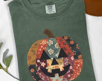 Puede incluir: Camiseta verde musgo con un diseño de calabaza de patchwork. La calabaza presenta telas estampadas florales en tonos naranja, rojo y marrón, con una cara negra. La camiseta es de la marca Comfort Colors.