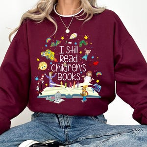 Può includere: Felpa bordeaux con la scritta "I still read children's books" e illustrazioni colorate di personaggi di libri per bambini. Il design include un libro, pastelli e vari personaggi dei cartoni animati.