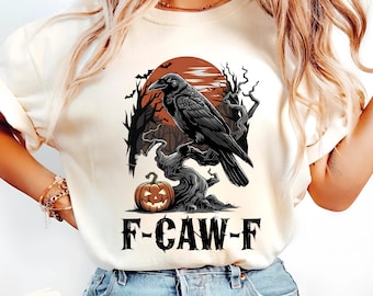 Pode incluir: Camiseta cor marfim com um design de Halloween. Apresenta um corvo preto pousado em um galho retorcido, uma abóbora esculpida e o texto "F-CAW-F". O fundo mostra um pôr do sol vermelho e silhuetas de árvores.