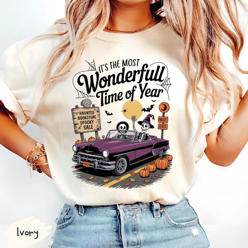 Puede incluir: Camiseta de color marfil con un dise&ntilde;o de Halloween. El dise&ntilde;o incluye un descapotable morado con esqueletos al volante, calabazas, murci&eacute;lagos y el texto "IT'S THE MOST Wonderfull Time of Year". Un letrero dice "Haunted Bookstore Spooky Sale."