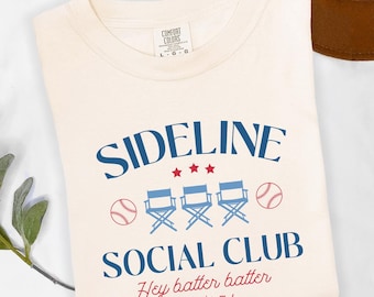 Op de afbeelding: Een ivoorwit T-shirt met een blauwe en rode afbeelding waarop "SIDELINE SOCIAL CLUB" staat met drie regisseursstoelen en honkballen. De tekst eronder luidt "Hey batter batter swing!"