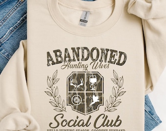 Puede incluir: Sudadera color crema con el texto "ABANDONED Hunting Wives Social Club" en un diseño de camuflaje. El diseño incluye un escudo con iconos relacionados con la caza y la frase "HELLO HUNTING SEASON. GOODBYE HUSBAND."