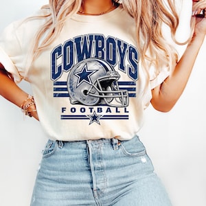 Könnte beinhalten: Ein cremefarbenes T-Shirt mit einem dunkelblauen Grafikdruck, der COWBOYS FOOTBALL zeigt. Das Design enthält eine detaillierte Abbildung eines Football-Helms mit einem Stern-Logo, zentriert zwischen horizontalen Streifen und einem Stern.