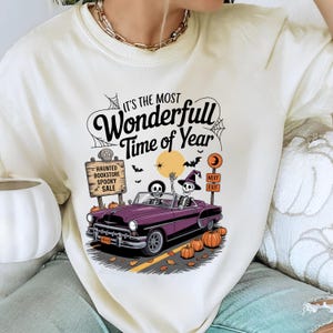 Puede incluir: Camiseta color crema con un gr&aacute;fico tem&aacute;tico de Halloween. El dise&ntilde;o presenta un descapotable morado con esqueletos al volante, calabazas y el texto "It's the Most Wonderfull Time of Year". Un letrero dice "Haunted Bookstore Spooky Sale".
