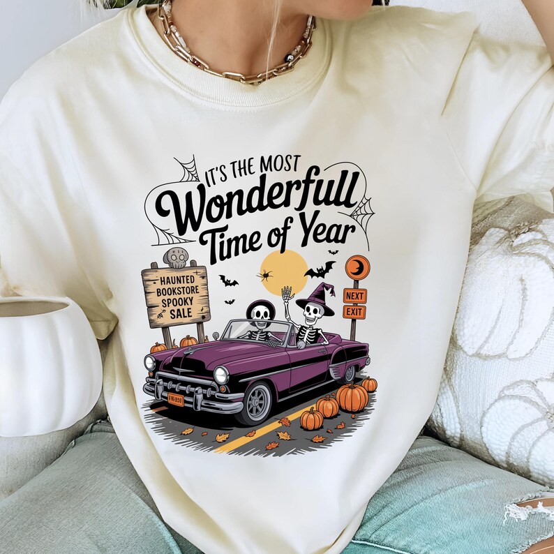 Puede incluir: Camiseta color crema con un gr&aacute;fico tem&aacute;tico de Halloween. El dise&ntilde;o presenta un descapotable morado con esqueletos al volante, calabazas y el texto "It's the Most Wonderfull Time of Year". Un letrero dice "Haunted Bookstore Spooky Sale".