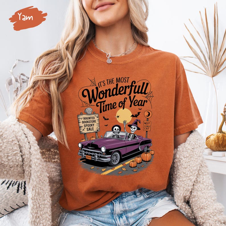 Puede incluir: Camiseta naranja con un dise&ntilde;o de Halloween. El dise&ntilde;o muestra un coche morado con esqueletos al volante, calabazas y el texto "It's the Most Wonderfull Time of Year". Un letrero dice "Haunted Bookstore Spooky Sale".