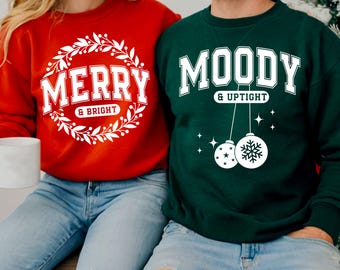 May include: Two sweatshirts, one red and one green. The red sweatshirt has the words "MERRY & BRIGHT" in white, surrounded by a wreath. The green sweatshirt says "MOODY & UPTIGHT" in white, with hanging ornament graphics. Both are casual wear.