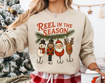 May include: Beige sweatshirt with the text "REEL IN THE SEASON" above four Christmas-themed fishing lures: a snowman, a nutcracker, Santa Claus, and a reindeer. The lures hang from a green pine branch.