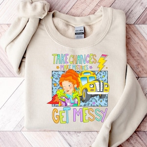 Op de afbeelding: Een beige sweater met ronde hals en een grafische print van een roodharig personage, een kleine groene draak en een gele schoolbus. De tekst luidt "TAKE CHANCES MAKE MISTAKES GET MESSY" met een illustratie van een geel bliksemschicht-potlood.