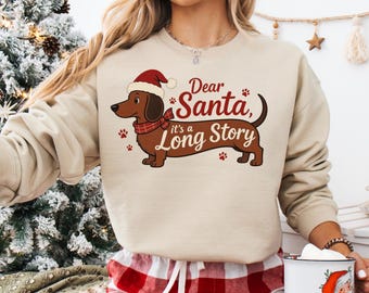 Peut inclure: Sweat-shirt beige avec un motif de Noël. Il présente un teckel portant un bonnet et une écharpe de Père Noël, avec le texte "Dear Santa, it's a Long Story". Des empreintes de pattes sont dispersées autour du chien, créant un thème de vacances.