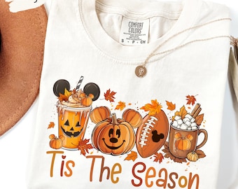 Può includere: T-shirt color avorio con grafica a tema autunnale. Il design include un pumpkin spice latte, una zucca di Mickey Mouse, un pallone da football americano e una tazza di cioccolata calda. Il testo "Tis The Season" è stampato sotto.